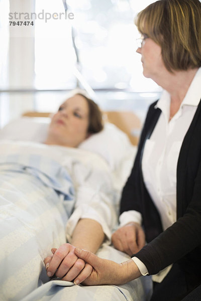 Seniorin mit der Hand der Tochter auf dem Bett liegend in der Krankenstation