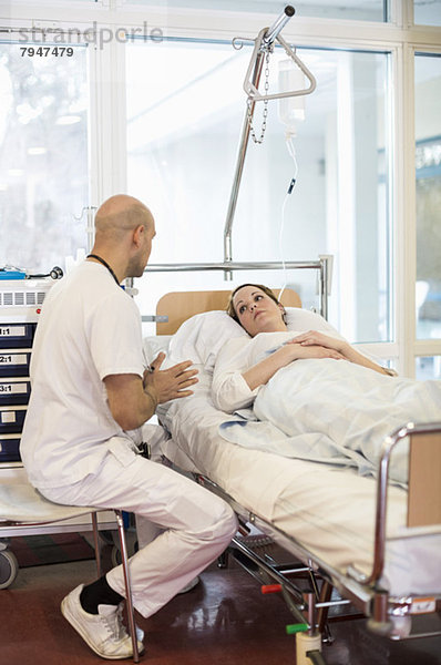 Mittlerer erwachsener männlicher Arzt  der mit dem auf dem Bett liegenden Patienten auf der Krankenstation kommuniziert.