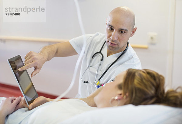 Mittlerer erwachsener männlicher Arzt  der dem auf dem Bett liegenden Patienten auf der Krankenstation eine digitale Tablette zeigt.