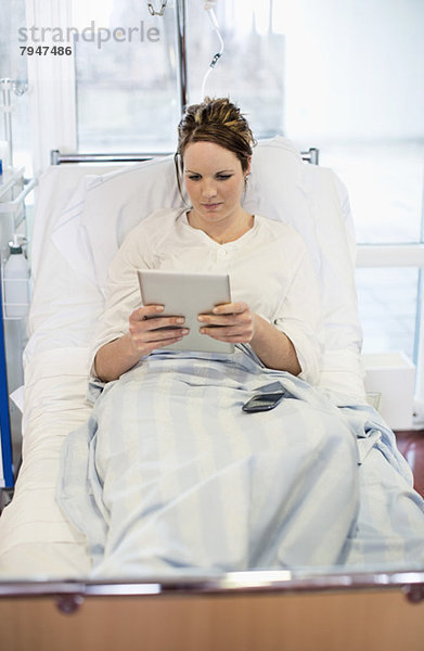 Mittlere erwachsene Patientin  die eine Tablette benutzt  während sie sich auf dem Bett in der Krankenstation entspannt.