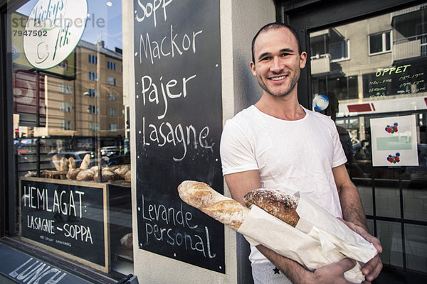 Porträt eines glücklichen jungen Mannes  der vor der Bäckerei steht und Brot hält.