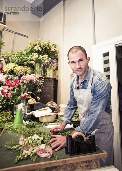 Porträt eines mittleren erwachsenen männlichen Floristen  der einen Blumenstrauß am Schreibtisch im Geschäft vorbereitet.