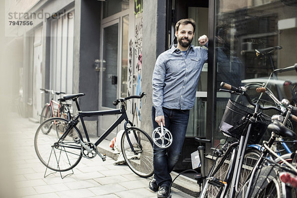 Porträt eines selbstbewussten mittleren erwachsenen Mannes mit Pedal am Eingang der Fahrradwerkstatt.