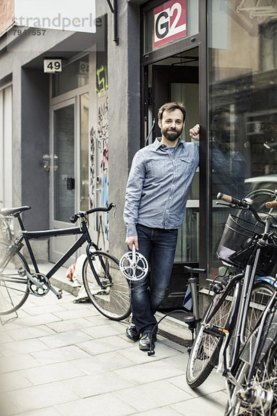 Porträt des mittleren erwachsenen männlichen Besitzers mit Pedal am Eingang der Fahrradwerkstatt