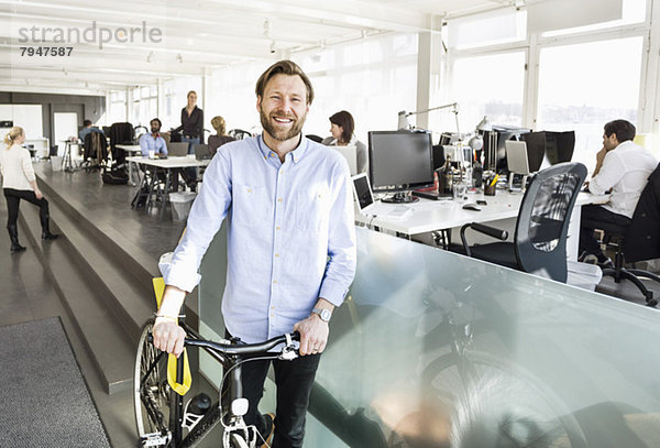 Porträt eines mittelständischen Geschäftsmannes  der mit Kollegen im Hintergrund im Büro Fahrrad hält