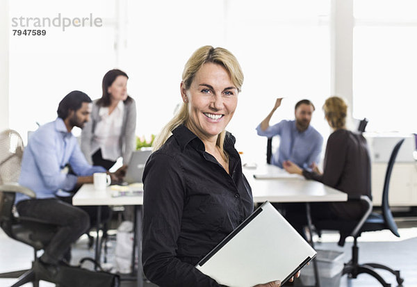 Porträt einer reifen Geschäftsfrau mit Laptop und Kollegen im Hintergrund im Büro