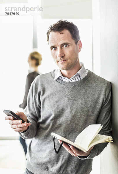 Porträt eines mittelständischen Geschäftsmannes mit Handy im Buch stehend im Büro