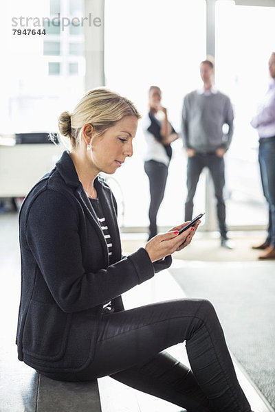 Seitenansicht der mittleren erwachsenen Geschäftsfrau per Handy mit Kollegen im Hintergrund im Büro