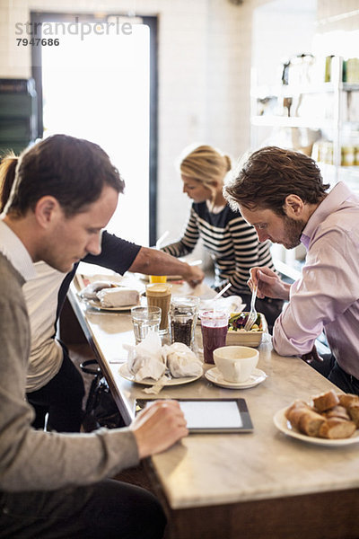 Mittlerer erwachsener Geschäftsmann mit digitalem Tablett und Kollegen beim Frühstück am Restauranttisch