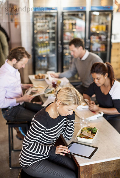 Großer Blickwinkel auf die mittlere erwachsene Geschäftsfrau mit digitalem Tablett und Kollegen im Hintergrund am Bürorestauranttisch