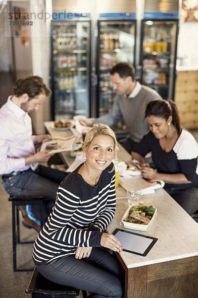 Hochwinkliges Porträt einer selbstbewussten Geschäftsfrau mit digitalem Tablett und Kollegen beim Frühstück im Hintergrund am Restauranttisch.