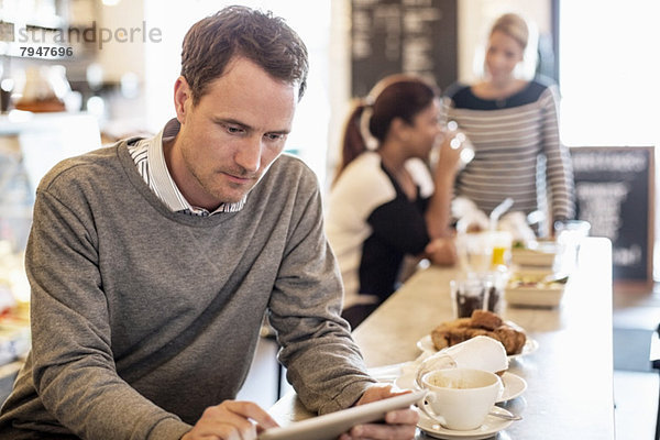 Mittlerer erwachsener Geschäftsmann mit digitalem Tablett am Restauranttisch mit Kollegen im Hintergrund