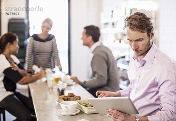 Mittlerer erwachsener Geschäftsmann mit digitalem Tablett am Restauranttisch mit Geschäftskollegen im Hintergrund