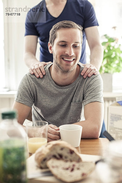 Fröhlicher schwuler Mann beim Kaffee  während der Partner hinter ihm am Tisch zu Hause steht.