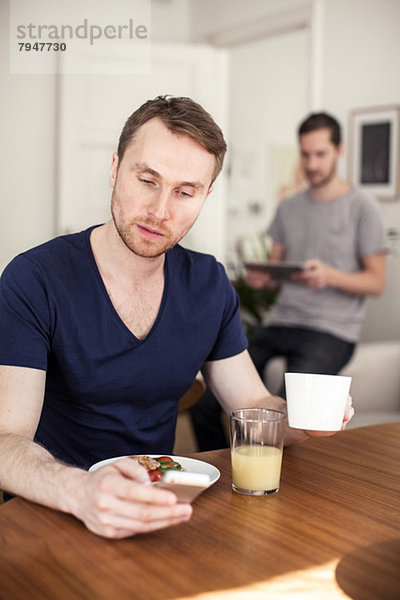 Junger schwuler Mann mit Handy beim Frühstück am Tisch mit Partner im Hintergrund