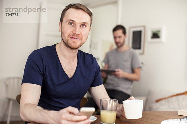 Porträt eines jungen schwulen Mannes mit Handy beim Frühstück am Tisch mit Partner im Hintergrund