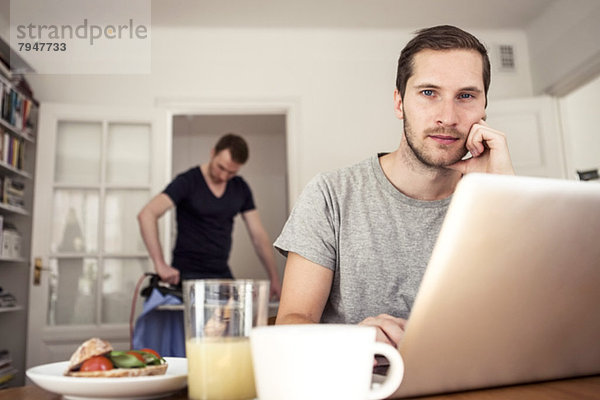 Porträt eines schwulen Mannes mit Laptop am Frühstückstisch mit Partnerbügeln im Hintergrund