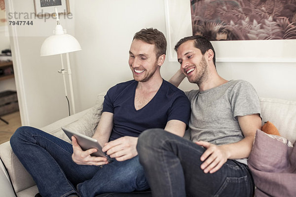 Fröhliches homosexuelles Paar  das zusammen auf dem Sofa zu Hause ein digitales Tablett benutzt.