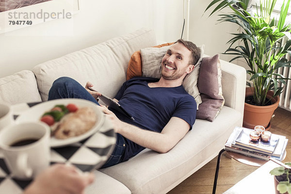 Abgeschnittenes Bild eines jungen schwulen Mannes mit Frühstück für den auf dem Sofa liegenden Partner.