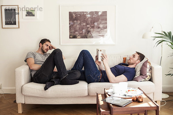 Junger schwuler Mann liest Buch  während der Partner ihn auf dem Sofa ansieht.