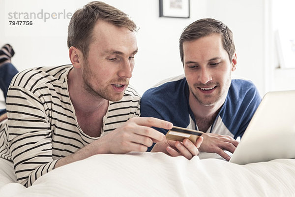 Homosexuelles Paar  das zusammen auf dem Laptop online einkauft  während es zu Hause im Bett liegt.