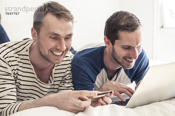 Glückliche homosexuelle Männer  die mit Kreditkarte und Laptop online einkaufen  während sie zu Hause im Bett liegen.
