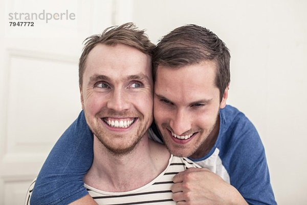 Fröhlicher junger schwuler Mann schaut weg  während der Partner zu Hause von hinten umarmt wird.