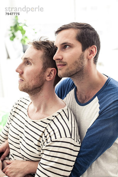 Nachdenkliches junges homosexuelles Paar schaut weg  während es sich zu Hause umarmt.