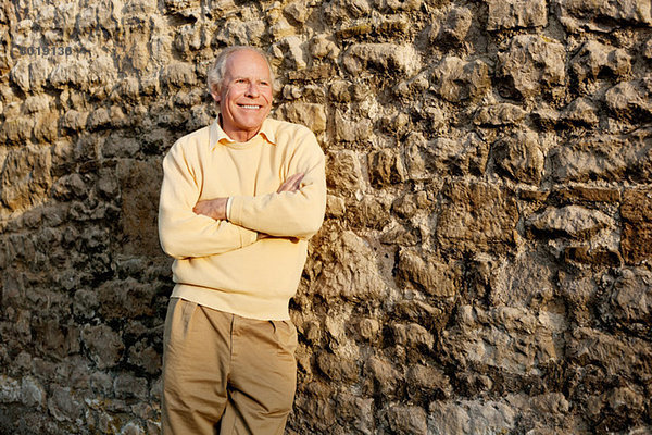 Älterer Mann  der sich mit gekreuzten Armen an die Steinmauer lehnt.