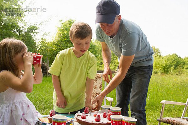Vater mit zwei Kindern beim Geburtstagskuchenschneiden im Freien