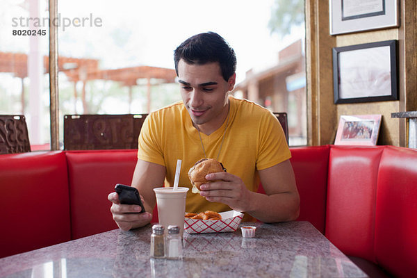 Junger Mann  der auf dem Handy schreibt und Fast Food im Diner isst.