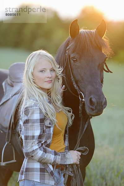 Junge Frau steht neben einem Pferd auf einer Wiese bei Sonnenuntergang