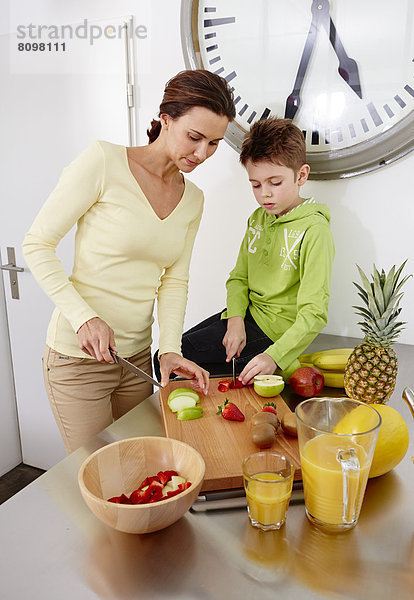 Mutter und Sohn schneiden verschiedene Sorten Obst auf Schneidebrett in Küche