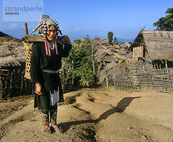 Akha-Frau in traditioneller Kleidung  mit Rückentrage  in einem Bergdorf mit Bambushütten