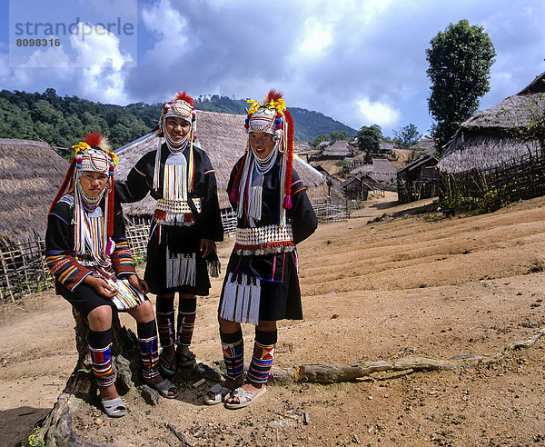 Drei Akha-Mädchen in einem Bergdorf mit traditioneller Kleidung und Kopfbedeckung  Bambushütten mit Strohdächer