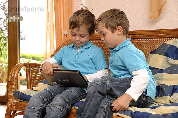 Zwei Jungen  Zwillinge  sitzen nebeneinander auf einer Korbbank im Wohnzimmer und spielen auf einem Tablet-PC