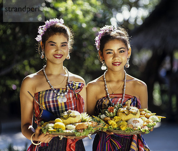 Zwei junge Frauen in traditionellen Kostümen mit geschnitzten exotischen Früchten  Rose Garden