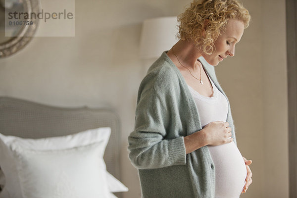 Lächelnde schwangere Frau berührt den Bauch im Schlafzimmer