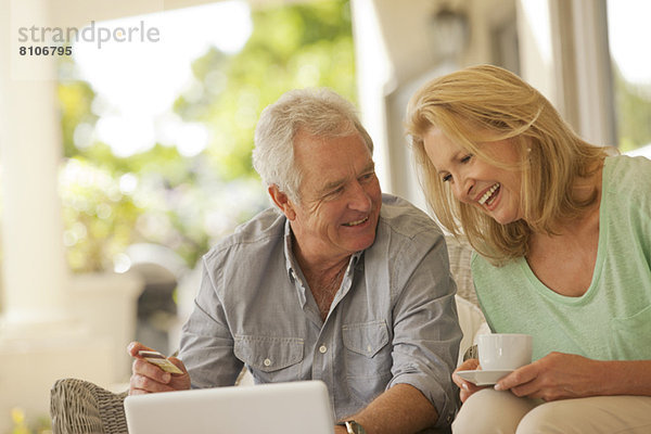 Lächelndes Paar trinkt Kaffee und kauft online auf der Terrasse ein.