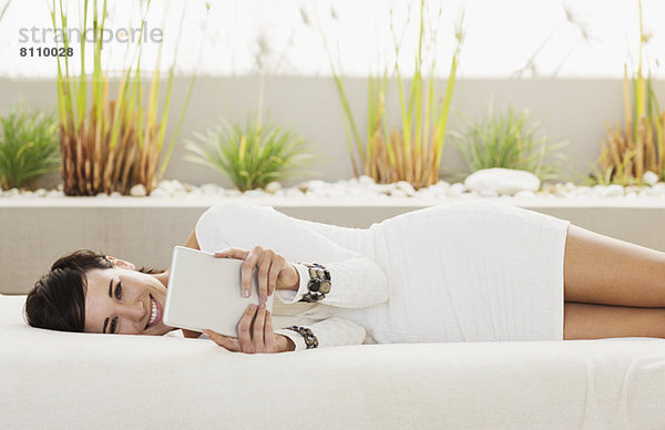 Porträt einer lächelnden Frau  die auf einem Sofa auf der Terrasse liegt und ein digitales Tablett benutzt.