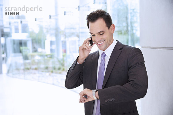 Lächelnder Geschäftsmann im Gespräch mit dem Handy und Blick auf die Armbanduhr