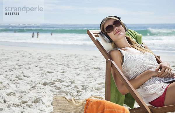 Gelassene Frau hört Kopfhörer im Liegestuhl am Strand