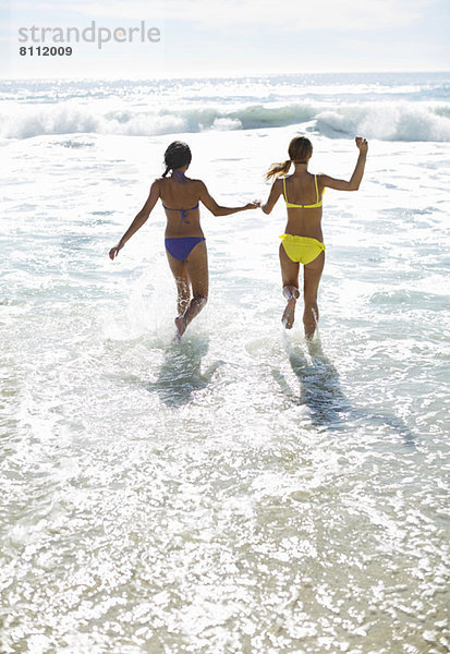 Freunde in Bikinis halten sich an den Händen und laufen ins Meer.