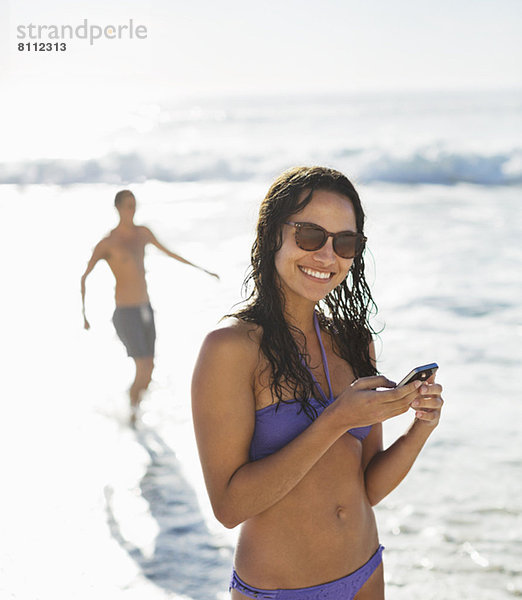 Porträt der lächelnden Frau im Bikini mit Handy am Strand
