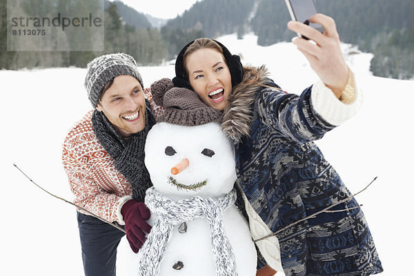 Enthusiastisches Paar beim Selbstporträt mit Schneemann im Schneefeld