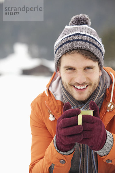 Nahaufnahme des lächelnden Mannes mit Strickmütze und Handschuhen beim Kaffeetrinken im Schneefeld