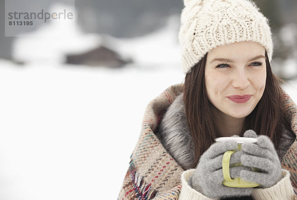 Nahaufnahme einer lächelnden Frau mit Strickmütze und Handschuhen beim Kaffeetrinken im Schneefeld