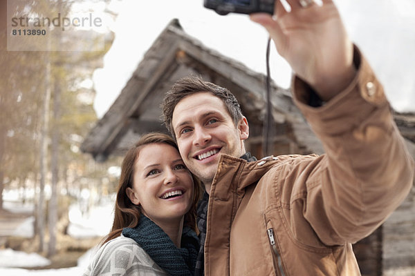 Lächelndes Paar beim Selbstporträt mit Digitalkamera außerhalb der Schneekabine