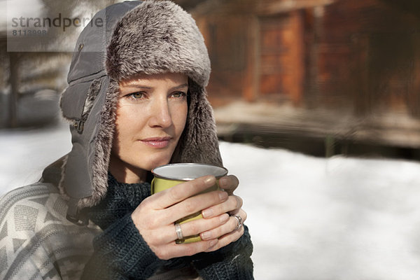 Nachdenkliche Frau mit Pelzmütze trinkt Kaffee außerhalb der Kabine