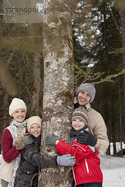 Porträt einer glücklichen Familie  die den Baumstamm in verschneiten Wäldern umarmt.